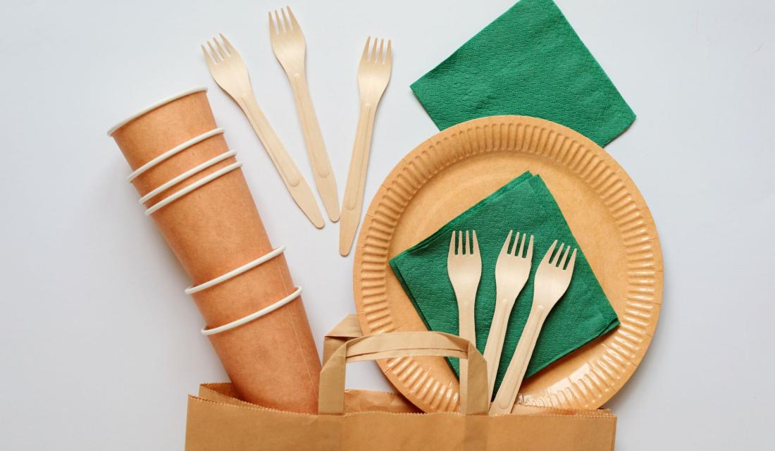 Becher, Teller und Servietten aus Papier und Besteck aus Holz über einer Papiertüte