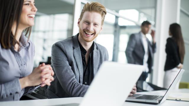 Mann im Büro vor Laptop lacht mit Kollegin