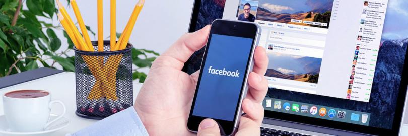 Mann am Schreibtisch hält Smartphone mit dem Schriftzug facebook auf dem Display in seinen Händen, dahinter ein aufgeklappter Laptop mit geöffneter Facebook-Seite