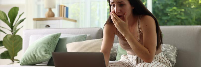 Junge Frau sitzt in schockierter Haltung vor einem Laptop bei sich zu Hause im Wohnzimmer