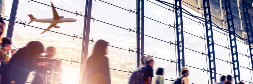 in geschäftiges Flughafenterminal mit Passagieren in Bewegung, die als verschwommene Silhouetten vor einer großen Glasfassade dargestellt sind. Im Hintergrund ist ein Flugzeug beim Start durch das Fenster zu sehen, während helles Sonnenlicht den Raum flutet und lange Schatten sowie Lichtreflexe auf dem glänzenden Boden erzeugt.