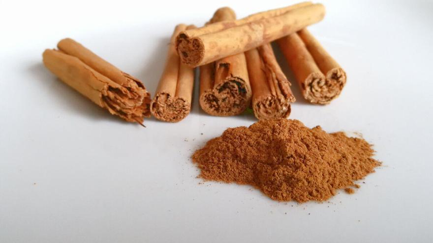 Sechs Cassia-Zimtstangen liegen auf einer weißen Fläche; davor ein Häufchen gemahlener Zimt