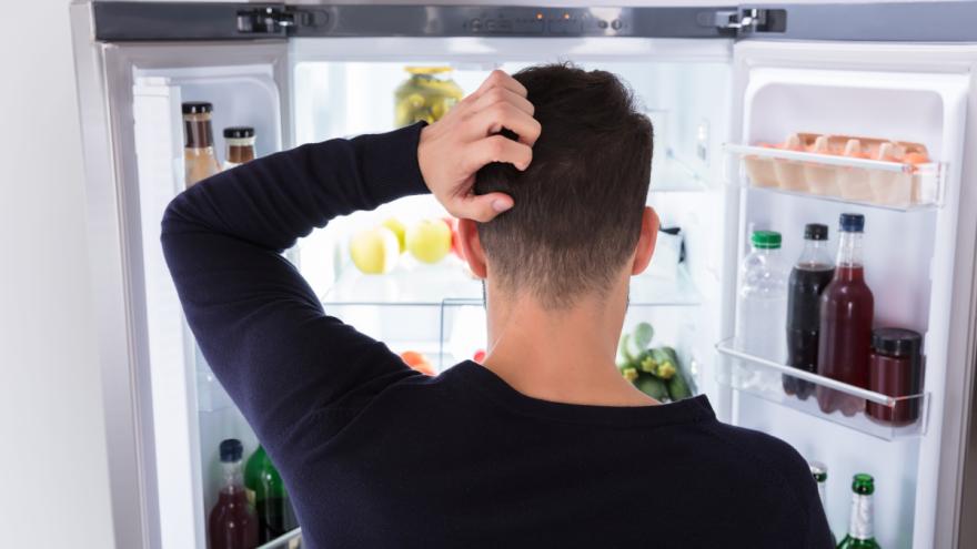 Ein Mann guckt in einen geöffneten Kühlschrank und kratzt sich dabei am Kopf.