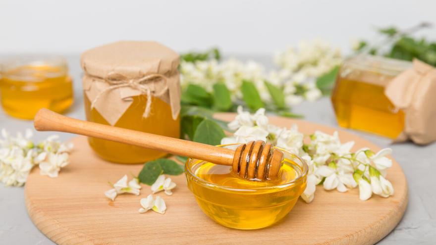 Honigglas, Schüssel mit Honig und Honiglöffel auf Holzbrett, umgeben von Blumen
