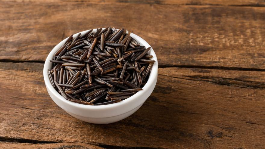 Eine kleine weiße Schüssel mit rohem Wildreis steht auf einem Holztisch.