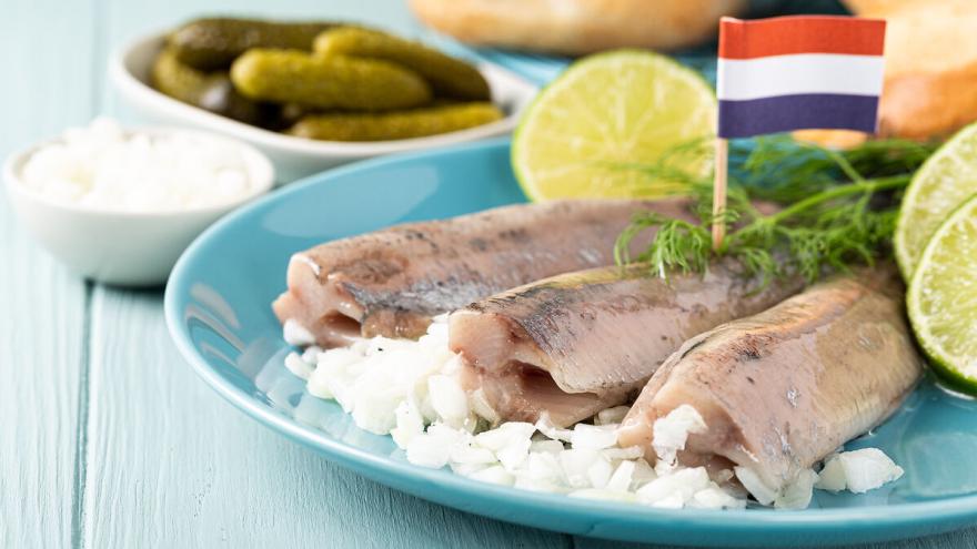 Holländischer Matjes auf einem blauen Teller, daneben Zitronenscheiben, Zwiebelstückchen und Kräuter