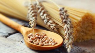 Rohe Spaghetti liegen auf einem Holztisch, darauf eine Weizenähre, daneben ein Holzlöffel mit Weizenkörnern.