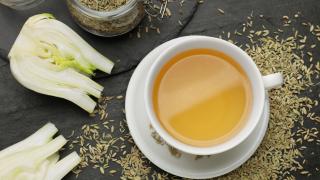 Eine Tasse Fencheltee steht auf einem Tisch, daneben zwei Hälften einer Fenchelknolle und Fenchelsamen.