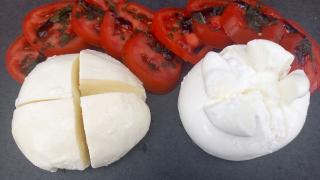 Auf einer schwarzen Arbeitsplatte liegen je ein Stück Mozzarella und ein Stück Burrata, darüber in Scheiben aufgeschnittene Tomaten.
