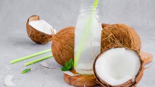 Mehrere Kokosnüsse, eine davon aufgeschlagen, liegen auf einem Tisch. In der Mitte steht eine Glasflasche mit Strohhalm, befüllt mit Kokoswasser.