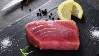 Ein rohes Stück Thunfisch liegt auf einem schwarzen Brett, daneben eine Zitronenscheibe, schwarze Pfefferkörnern und ein Zweig Rosmarin.