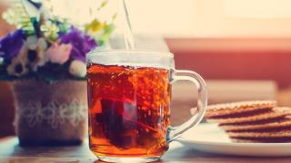 Eine Tasse Tee steht auf einem Tisch, links davon stehen Blumen, rechts davon ein Teller mit Keksen