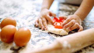 Kinderhände stechen mit einem Teigausstecher eine Form aus einem rohen Teig. Daneben liegen drei rohe Eier und ein Teigroller..
