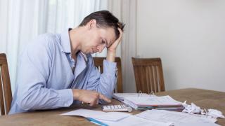 Mann schaut verzweifelt auf einen Taschenrechner. Um ihn herum auf dem Tisch liegen Aktenordner.