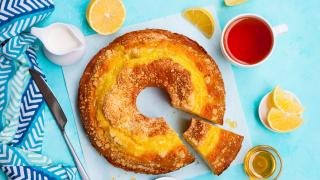 Ein Rührkuchen von oben, daneben befinden sich eine aufgeschnittene Zitrone, eine Tasse Tee, ein Kännchen Milch, ein Messer und ein Löffel.