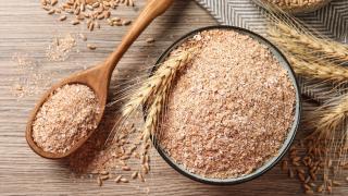 Eine Glasschüssel mit Weizenkleie steht auf dem Tisch. Daneben liegt ein Holzlöffel, der ebenfalls mit Weizenkleie gefüllt ist.