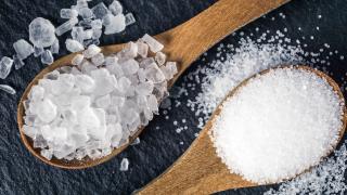 Zwei Holzlöffel je mit grobem und feinem Salz liegen auf einer schwarzer Granitplatte, umgeben von verstreuten Salzkörnern.