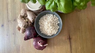 Eine Schüssel mit Pankomehl steht auf einem Holztisch. Daneben rote Zwiebeln, Ingwer und ein Basilikumstock.