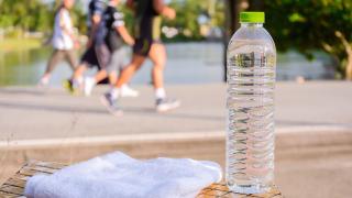 Im Vordergrund steht eine Wasserfasche aus PET, daneben liegt ein weißes Handtuch. Im Hintergrund joggt eine kleine Gruppe Läufer.