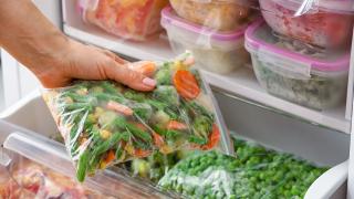 Eine Hand holt aus einem geöffnetem Kühlschrank eine Tüte mit gefrorenem Gemüse heraus.