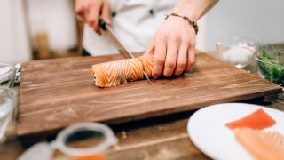 Eine mit Lachsscheiben ummantelte Sushirolle liegt auf einem Holzbrett. Sie wird von einer Hand gehalten, die andere Hand hält ein großes Messer, mit dem sie die Rolle in der Mitte durchschneidet.