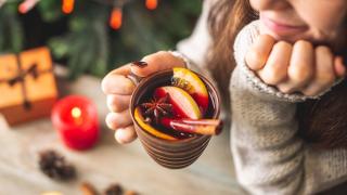 Eine Frauenhand hält eine Tasse mit einem winterlichen Getränk. Im Getränk befinden sich Orangenscheiben, Zimtstangen und Sternanis.