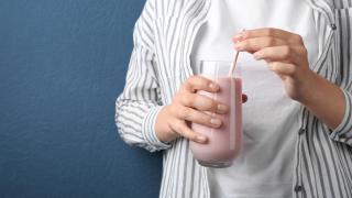 Eine Hand hält ein Glas mit einer hellrosa Trinkmahlzeit, drei Finger der anderen Hand umschließen den Strohhalm, der im Getränk steckt.