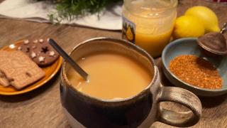 Eine Tasse hellbrauner Tee mit Löffel steht auf einem Holztisch, umgeben von Honig, Zitronen, losen Teeblättern und einem Teller mit Weihnachtsgebäck.